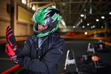 Foto de Conductor de deportes de motor afroamericano en casco con guantes y de pie cerca del circuito, go-kart - Imagen libre de derechos