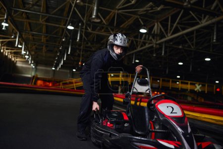 motorsport and speed drive, focused kart driver in sportswear and helmet pushing go kart on circuit