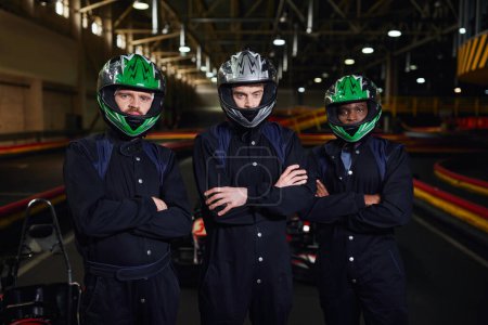 three confident go kart drivers in sportswear and helmets standing with folded arms on circuit