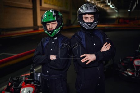 dos competidores de karts seguros en ropa deportiva y cascos de pie con los brazos cruzados en el circuito