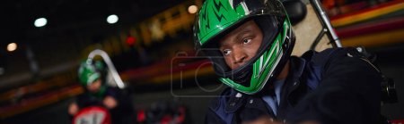 africano americano hombre en casco conducción ir kart en circuito cerca amigo en borrosa telón de fondo, bandera