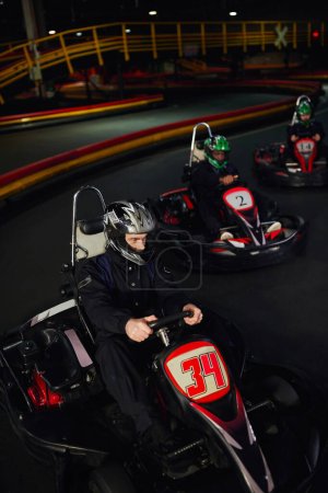 Foto de Conductores diversos enfocados en cascos que conducen el kart del go en circuito cubierto, automovilismo y adrenalina - Imagen libre de derechos