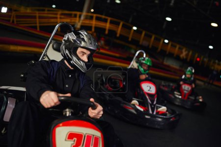 Foto de Hombre concentrado conduciendo ir kart cerca de diversos conductores en cascos en circuito interior, adrenalina - Imagen libre de derechos