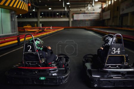 two diverse racers in helmets and sportswear driving go kart on indoor circuit, competitors