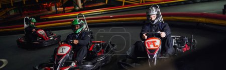 three multicultural men in helmets and sportswear driving go kart on indoor circuit, banner