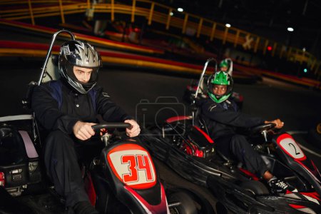 Foto de Competidores multiculturales conducción ir kart en circuito cubierto, carreras de velocidad y automovilismo - Imagen libre de derechos