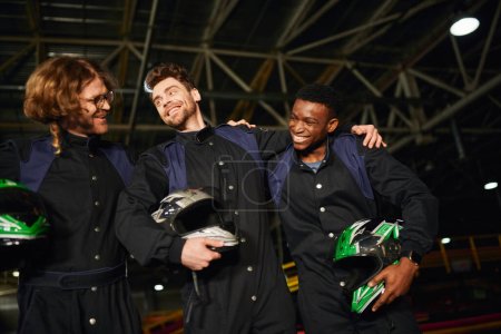 team of interracial and happy go kart drivers in protective suits hugging and holding helmets