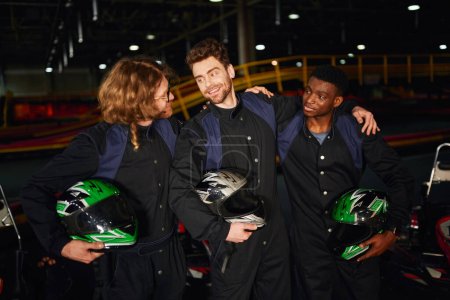 Gruppe gemischtrassiger und glücklicher Go-Kart-Fahrer in Schutzanzügen, die sich umarmen und Helme halten