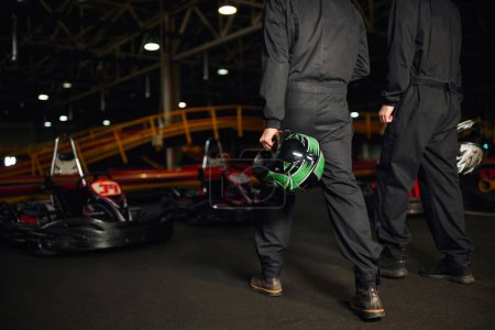 vista recortada de los conductores de karts go en ropa deportiva caminando en el circuito y la celebración de cascos, competidores