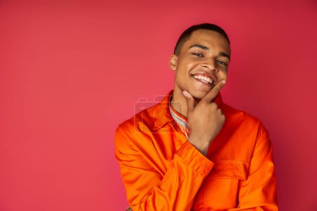 happy african american man in orange shirt touching face and looking away on red background