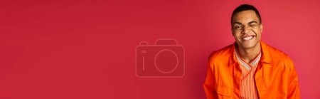 homme afro-américain souriant en chemise orange clin d'oeil à la caméra sur fond rouge, bannière, espace de copie