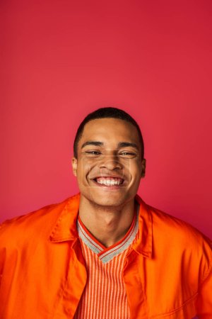 optimista hombre afroamericano sonriendo a la cámara sobre fondo rojo, camisa naranja, retrato