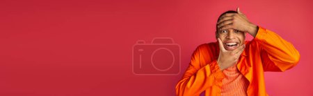 playful african american man looking at camera and touching face, red background, banner, copy space