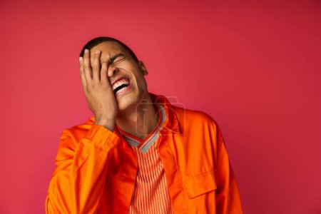 afrikanisch-amerikanisches verschleierndes Gesicht und Lachen, Aufregung, orangefarbenes Hemd, trendiger roter Hintergrund