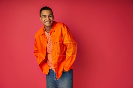 trendy african american man with hands in pocket smiling at camera on red background