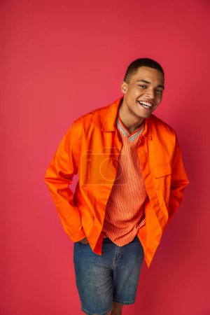 joyful african american guy in orange shirt, with hands in pockets, smiling at camera on red