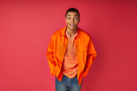 Téléchargez les photos : Surpris homme afro-américain avec les mains dans la poche, en chemise orange regardant la caméra sur rouge - en image libre de droit