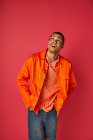 Photo for Positive and curious african american in orange shirt looking up on red background, hands in pockets - Royalty Free Image