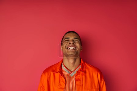 überglücklicher afrikanisch-amerikanischer Mann in orangefarbenem Hemd lacht mit geschlossenen Augen auf rot, Aufregung