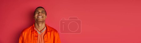 excited african american guy laughing with closed eyes on red, orange shirt, banner, copy space
