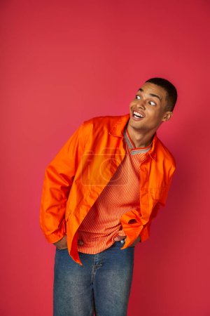 erstaunt afrikanisch-amerikanischer Mann lächelt und schaut weg auf rot, Hände in den Taschen, orangefarbenes Hemd