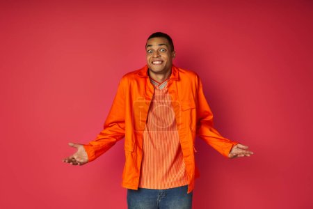 discouraged african american man in orange shirt showing shrug gesture on red background