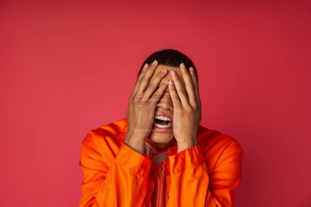 Photo for Young depressed african american man in orange shirt obscuring face with hands on red background - Royalty Free Image