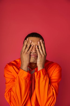 Foto de Afro-americano con problemas en camisa naranja que oscurece la cara con las manos sobre el fondo rojo - Imagen libre de derechos