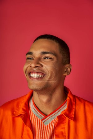 Téléchargez les photos : Portrait d'un jeune homme afro-américain avec un sourire radieux, chemise orange élégante, fond rouge - en image libre de droit