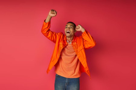 aufgeregter afrikanisch-amerikanischer Typ in orangefarbenem Hemd zeigt Erfolgsgeste und schreit auf rotem Hintergrund