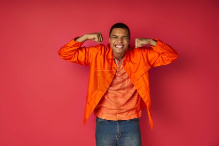 Téléchargez les photos : Homme afro-américain expressif en chemise orange démontrant la force et regardant la caméra sur rouge - en image libre de droit
