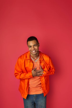 glad african american man touching chest, smiling at camera on red, grateful gesture, orange shirt