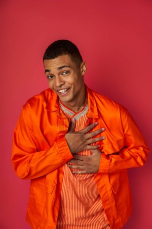 dankbarer und lächelnder afrikanisch-amerikanischer Mann in orangefarbenem Hemd, der die Brust berührt und in die Kamera auf rot blickt