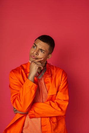 deep in thought african american man in orange shirt touching chest and looking away on red