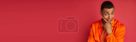 Foto de Hombre afroamericano pensativo y tenso en camisa naranja mirando a la cámara en rojo, pancarta, espacio de copia - Imagen libre de derechos