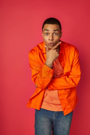 Foto de Hombre afroamericano emocionado en camisa naranja, con la mano cerca de la cara mirando a la cámara en rojo - Imagen libre de derechos