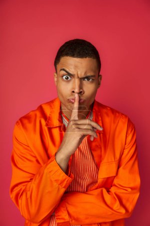 strict african american man in orange shirt looking at camera and showing hush gesture on red