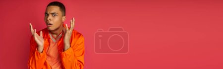 schockierter Afroamerikaner in orangefarbenem Hemd gestikuliert und blickt in die Kamera auf rot, Banner, Kopierraum