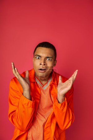 discouraged african american man in orange shirt gesturing and looking at camera on red