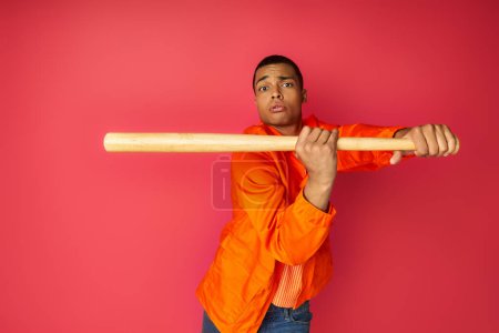 konzentrierter afrikanisch-amerikanischer Mann in orangefarbenem Hemd mit Baseballschläger und roter Kamera