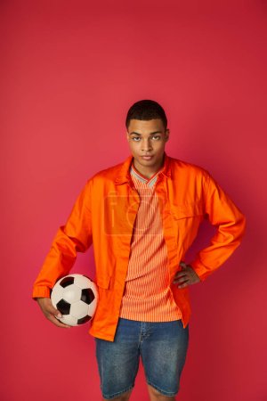 confiant afro-américain en chemise orange debout avec ballon de football et la main sur la hanche sur rouge
