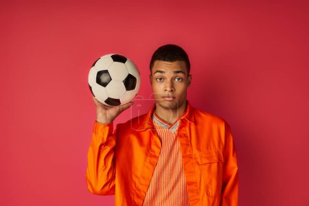 youthful african american guy in orange shirt holding soccer ball and looking at camera on red