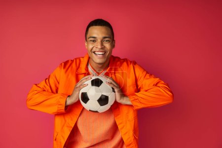 fröhlicher afrikanisch-amerikanischer Mann in orangefarbenem Hemd, Fußballball in der Hand und lächelt in die Kamera auf rot