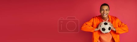 Photo for Happy african american man in orange shirt holding soccer ball on red, banner, copy space - Royalty Free Image