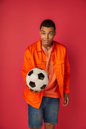 smiling and skeptical african american man in orange shirt showing soccer ball on red background