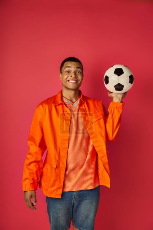 fröhlicher afrikanisch-amerikanischer Mann in orangefarbenem Hemd, Fußballball in der Hand und lächelnd in die Kamera auf rot