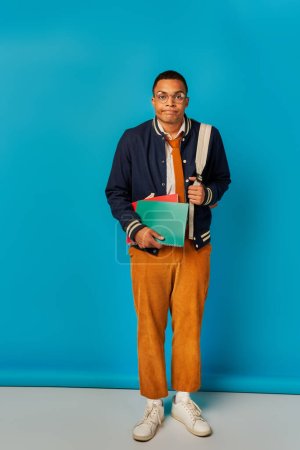 Téléchargez les photos : Ennui étudiant afro-américain avec cahiers et sac à dos joues gonflantes sur fond bleu - en image libre de droit