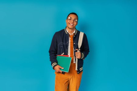 happy and trendy african american student with backpack and notebooks looking at camera on blue