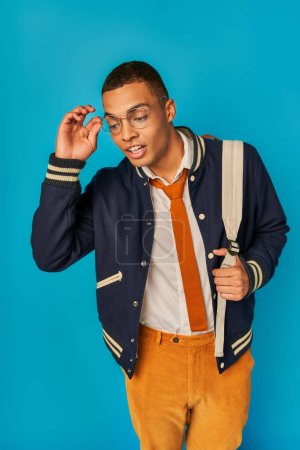 curious african american student adjusting eyeglasses and looking down on blue, notebooks, backpack