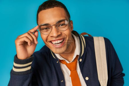 joyful african american student in jacket adjusting eyeglasses, looking at camera on blue, portrait